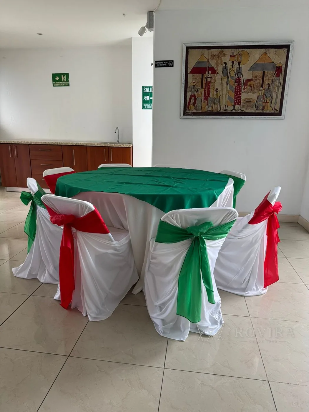 Mesa redonda con lazos en rojo y verde sobre forros blancos y cuadro típico costarricense de fondo