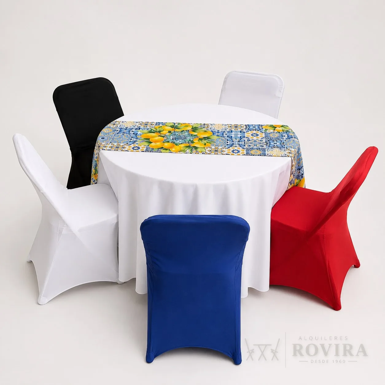 Forros elásticos (spandex) para silla en blanco, negro, azul rey y rojo alrededor de una mesa redonda con mantel blanco y camino de azulejos con limones, foto de producto