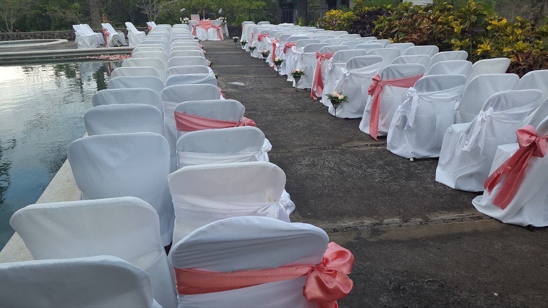 Boda junto a piscina al aire libre, sillas forradas blancas con lazo coral