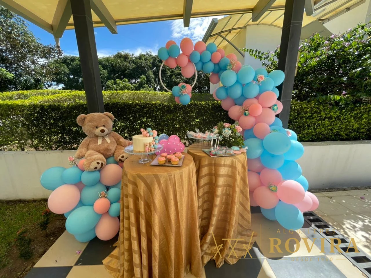 Mesa cocktailera alta con mantel dorado satinado y decoración de globos, en jardín