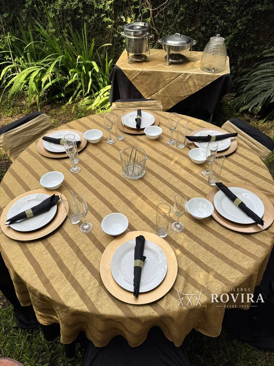 Mesa redonda con mantel a rayas doradas, vajilla completa y copas, mesa de servicio con chafing dishes al fondo en jardín