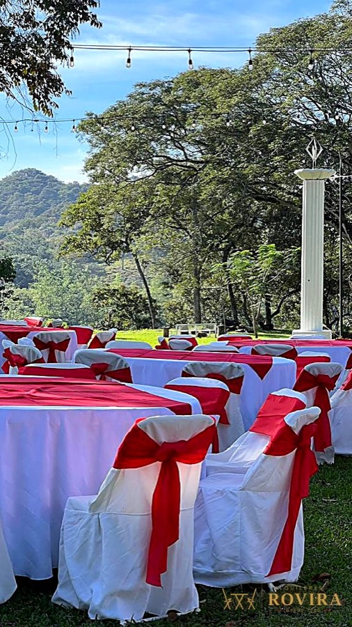 Recepción al aire libre con vista a la montaña, mesas redondas con mantel blanco y forros de silla con lazo rojo, luces guirnalda