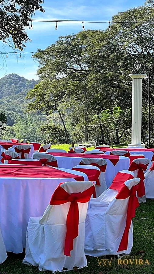 Recepción al aire libre con vista a la montaña, mesas redondas con mantel blanco y forros de silla con lazo rojo, luces guirnalda