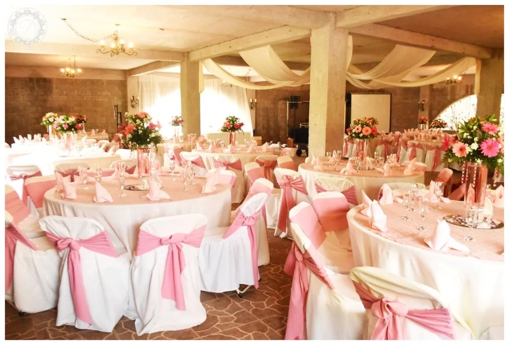 Salón de bodas con techos de tela blanca, mesas redondas vestidas en rosa y sillas con lazo rosa, arco floral colgante